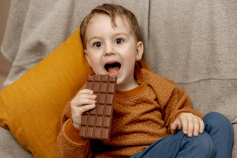 可爱的男孩坐着沙发上首页吃巧克力酒吧孩子糖果糖糖果孩子享受美味的甜点学前教育孩子休闲服装积极的情感