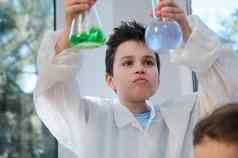 英俊的学校男孩持有玻璃实验室烧瓶化学物质看反应采取的地方化学类