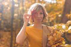 年轻的金发女郎女人覆盖眼睛红色的枫木叶秋天季节概念户外秋天女肖像特写镜头树叶
