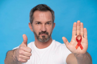 拇指手势成熟的男人。红色的丝带弓艾滋病意识象征棕榈穿白色t恤孤立的蓝色的背景现代医学医疗保健艾滋病意识概念