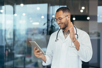 快乐的拉美裔医生医疗白色外套听诊器平板电脑电脑视频调用在线咨询病人男人。工作内部诊所办公室建筑