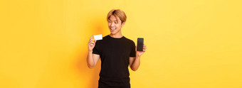 英俊的满意亚洲的家伙持有信贷卡显示智能<strong>手机屏幕</strong>微笑很高兴站黄色的背景
