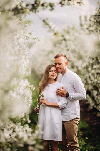 快乐年轻的夫妇爱站花园盛开的苹果树男人。白色衬衫女孩白色光衣服走开花公园