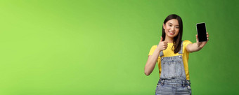 快乐的可爱的亚洲女人判断好智能手机游戏显示翘拇指标志扩展手臂持有电话屏幕现在太棒了应用程序促进应用程序站绿色背景