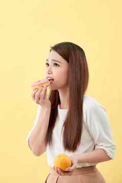 节食概念美丽的年轻的亚洲女人持有橙色甜甜圈黄色的背景选择垃圾食物健康的食物