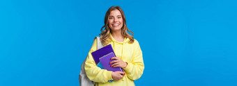 快乐的漂亮的金发碧眼的女孩微笑相机携带背包笔记本电脑论文研究学习语言课程站快乐的蓝色的背景