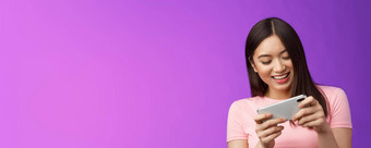 特写镜头快乐的有吸引力的亚洲女人浅黑肤色的女人有趣的花时间玩智能手机游戏笑微笑急切的赢得比赛持有电话水平打分数紫色的背景