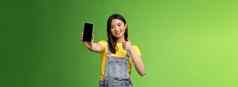 快乐的可爱的亚洲女人判断好智能手机游戏显示翘拇指标志扩展手臂持有电话屏幕现在太棒了应用程序促进应用程序站绿色背景