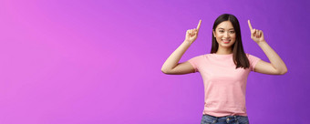 可爱的自信的黑头发的亚洲女人穿粉红色的t恤提高手显示<strong>促销</strong>指出手指咧着嘴笑邀请链接试演产品给方向站<strong>紫色</strong>的背景