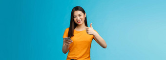 满意可爱的亚洲浅黑肤色的女人持有智能手机显示翘拇指很高兴推荐太棒了小工具给批准好应用程序同意朋友发布好博客文章站蓝色的背景