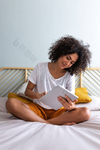年轻的多民族的女人坐着床上放松首页舒适的卧室垂直