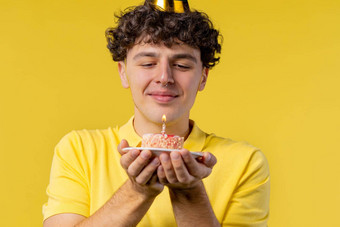 快乐生日男人。使吹蜡烛蛋糕卷曲的头发的的家伙笑着说庆祝周年纪念日年轻的少年黄色的背景