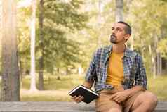 年轻的男人。坐着在户外持有书公园树背景知识信仰概念
