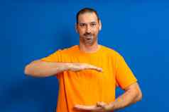 英俊的拉丁美洲人有胡子的男人。橙色t恤持有手孤立的蓝色的背景产品演讲