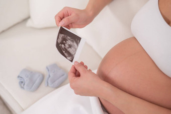 不知名的怀孕了女人坐在白色沙发持有超声波照片胎儿