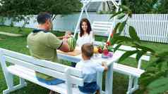年轻的家庭有趣的孩子们坐着晚餐表格花园夏天爸爸削减大西瓜治疗家庭午餐家庭
