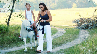 妈妈女儿走场女儿骑小马妈妈。持有小马缰绳快乐的快乐家庭假期在户外夏天森林