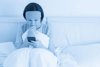 女人穿睡觉面具发短信智能手机坐在床上覆盖羽绒被