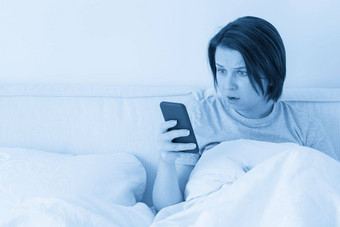 惊讶女人智能手机坐在床上覆盖羽绒被