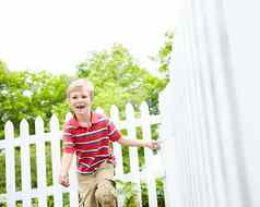 年轻的可爱的年轻的男孩运行坚持白色哨栅栏后院