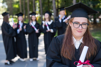 肖像年轻的高加索人女人眼镜研究生礼服背景同学们集团研究生学生在户外