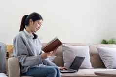 年轻的美丽的亚洲女人学生坐着沙发阅读书首页移动PC在线学习
