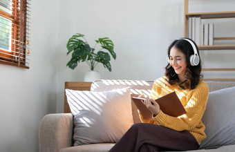 年轻的亚洲女人听音乐耳机写作请注意工作的想法日记书坐着灰色沙发生活房间
