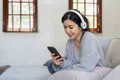 亚洲女人听音乐移动电话耳机平静首页周末放松坐沙发享受最喜欢的调优受欢迎的音乐现代无线耳机小工具生活房间