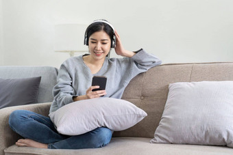 亚洲女人听音乐移动电话耳机平静首<strong>页</strong>周末放松坐沙发享受最喜欢的调优受<strong>欢迎</strong>的音乐现代无线耳机小工具生活房间