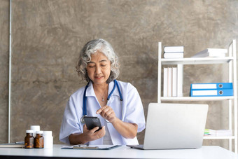医生高级头发灰白的女人白色医疗外套智能手机微笑坐移动PC办公室
