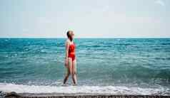 年轻的快乐女人走无忧无虑的海边快乐夫人红色的泳装金发碧眼的头发走海滨海滩穿太阳镜红色的紧身衣裤享受明亮的太阳射线海海洋海滩