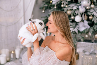 女人持有白色兔子象征一年特写镜头美丽的年轻的金发女郎女人持有兔子闪亮的衣服坐在圣诞节装饰房间