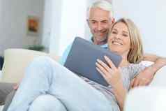 成熟的夫妇平板电脑放松沙发房子首页生活房间社会媒体互联网应用程序快乐微笑退休男人。女人数字技术视频调用电影流媒体新闻