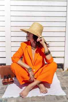 时尚的女人橙色西装他坐在地毯白色条纹背景手珠宝环手镯凉鞋袋站一边一边