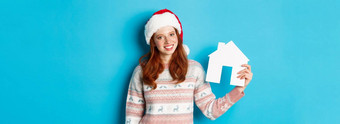 假期广告片真正的房地产概念快乐的红色头发的人女人圣诞老人他持有纸房子手微笑站毛衣蓝色的背景