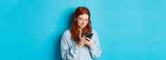 年轻的十几岁的红色头发的人女孩阅读消息智能手机微笑移动电话站蓝色的背景