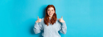 快乐的青少年女孩红色的头发显示拇指批准赞美手势站蓝色的背景