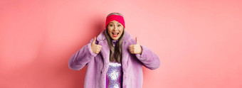 时尚购物概念快乐的亚洲女人显示竖起大拇指批准推荐促销活动站时尚的<strong>紫色</strong>的皮毛<strong>外套</strong>粉红色的背景