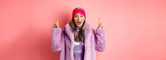 快乐上了年纪的亚洲夫人指出手指惊讶相机显示很酷的促销提供站粉红色的背景紫色的假的皮毛外套