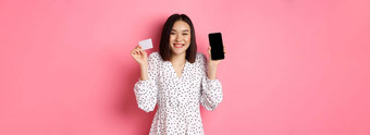 快乐的朝鲜文女人显示智能手机屏幕信贷卡支付互联网订单展示在线购物应用程序站粉红色的背景