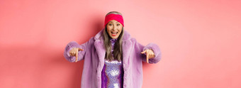 时尚购物概念快乐朝鲜文高级女人指出手指显示太棒了促销交易穿时尚的紫色的皮毛外套头巾粉红色的背景