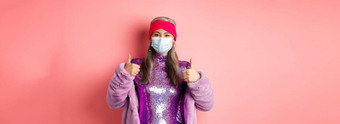 冠状病毒购物概念时尚的亚洲祖母呼吸器闪闪发光的紫色的衣服人造皮毛外套显示竖起大拇指推荐商店脸面具