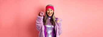 快乐的亚洲高级女人灰色头发穿聚会，派对闪闪发光的衣服显示美味的甜甜圈微笑快乐吃甜蜜的食物站粉红色的背景