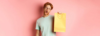 有趣的英俊的男人。红色的头发穿眼镜t恤持有黄色的购物袋购买礼物促销提供站粉红色的背景