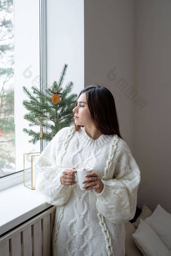 女人温暖的白色冬天毛衣说谎床上首页圣诞节夏娃持有杯棉花糖冷杉树