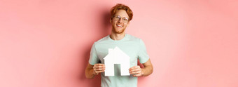 真正的房地产快乐的年轻的男人。红色的头发穿眼镜t恤显示纸房子断路微笑购买公寓粉红色的背景