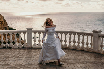 浪漫的新娘金发碧眼的女孩白色婚礼衣服开放肩膀摆姿势开放阳台背景海岩石时尚的年轻的女人站阳台海洋日落