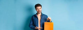 英俊的高加索人男人。显示橙色购物袋微笑购买礼物站蓝色的背景