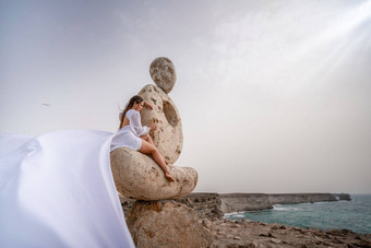 女人坐在石头雕塑使<strong>大石头</strong>穿着白色长衣服背景海天空衣服发展风