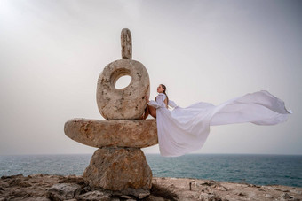 女人坐在石头<strong>雕塑</strong>使大石头穿着白色长衣服背景海天空衣服发<strong>展</strong>风